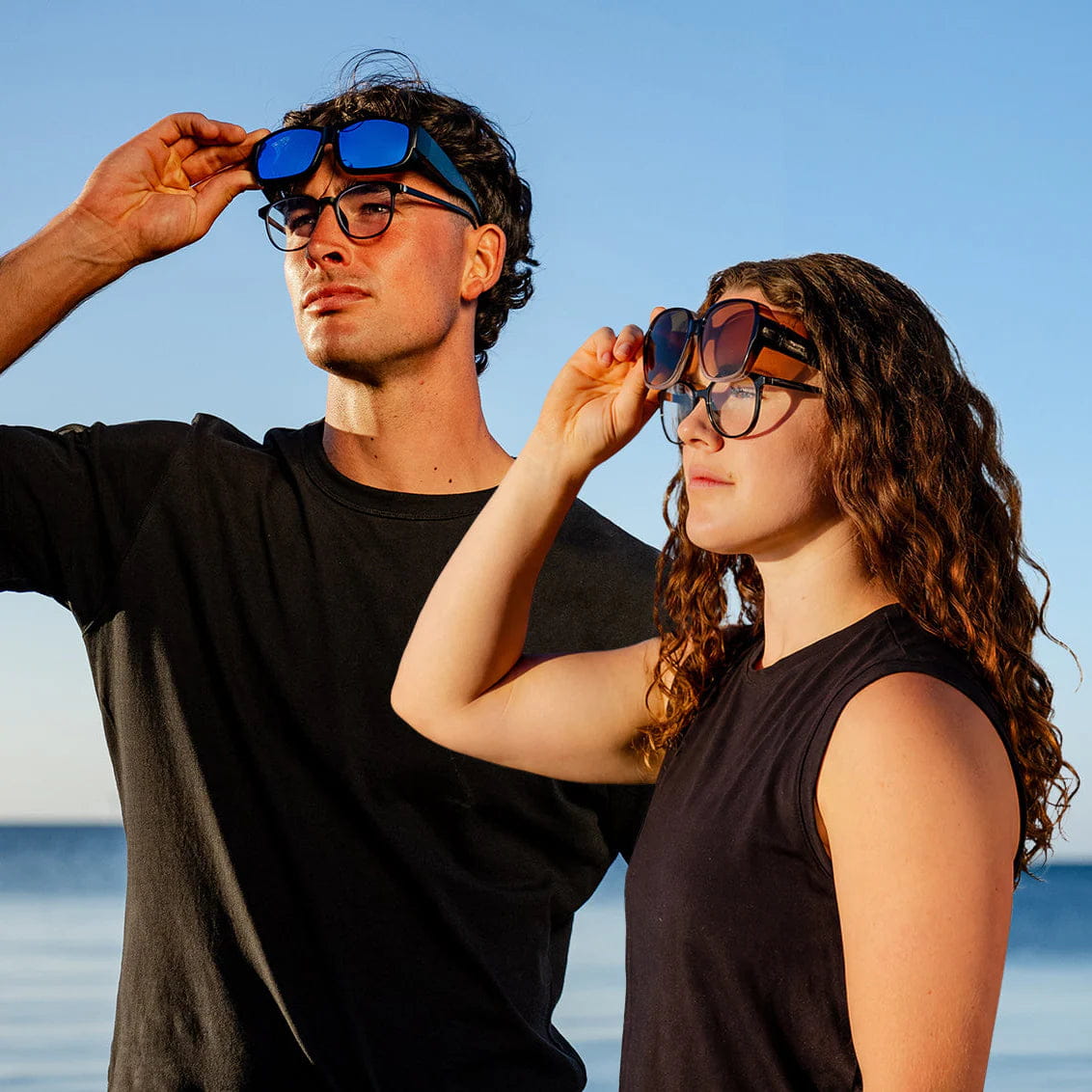 A man and a woman wearing fit-over sunglasses over their prescription glasses by the sea
